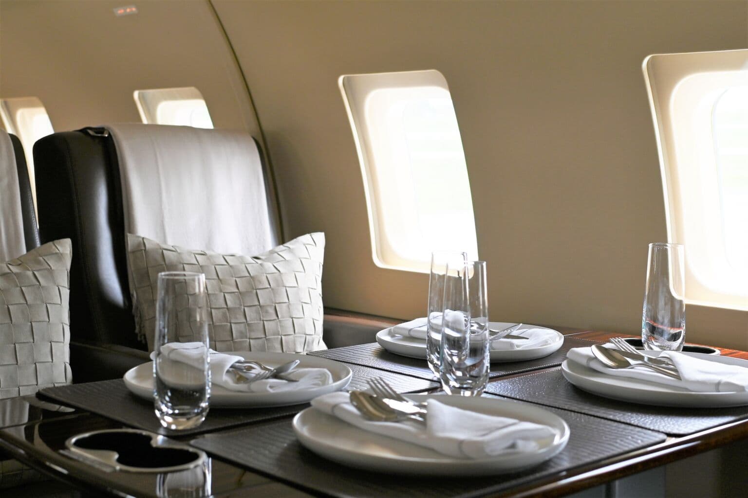 A private jet cabin table laid for a meal, with white plates, folded napkins and cut-glass tumblers beside a sunlit window.