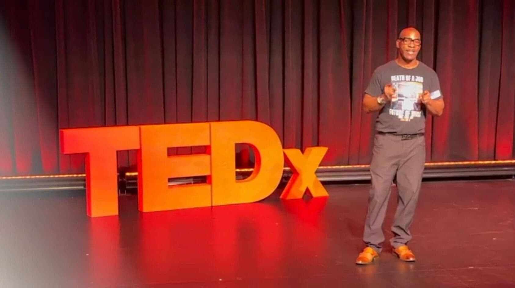 Mike Johns speaking on stage at a TEDx event, holding a copy of his book Death of a Job.